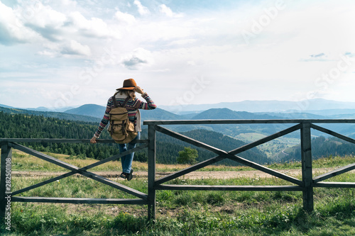 Stylish hipster girl in hat sit on fence and looking on top of mountains. Happy woman with backpack exploring summer mountains. Travel and wanderlust concept. Amazing atmospheric moment.