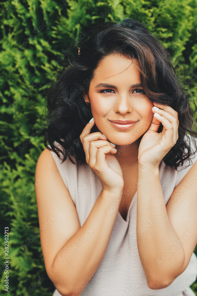 Outdoor portrait of beautiful young woman with professional makeup and hair styling, leaning on green natural background Outdoor portrait of beautiful young woman with professional makeup and hair styling, leaning on green natural background