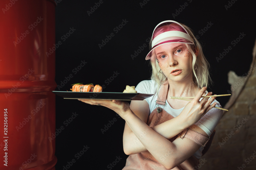 Portrait of attractive lady with makeup and pink cap holding a plate with sushi rolls and chopsticks looking into the camera with a serious face. Japanese food and fashion girl, fashion photo.