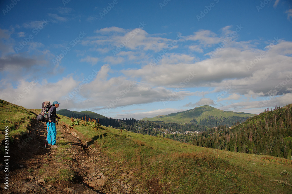 Obraz premium Man standing on the dirt road with hiking backpack and sticks in the background of tree covered hills
