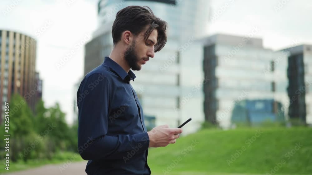 Zoom in side view of bearded young businessman in black shirt text messaging on cell phone and looking away thoughtfully while standing on city street, slow motion