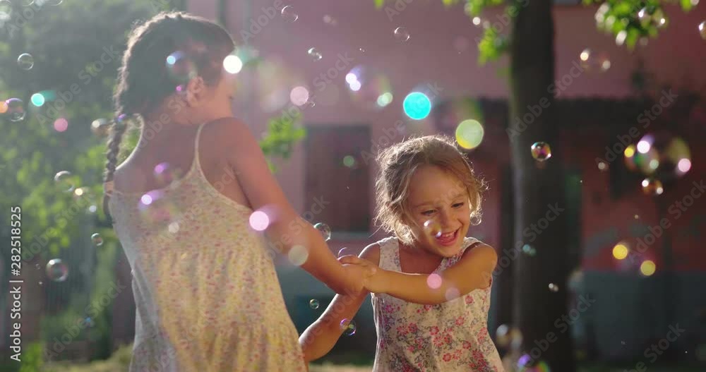 Authentic shot of two cute little girls sisters having fun to dance and playing with soap bubbles outside their house on a sunny day.