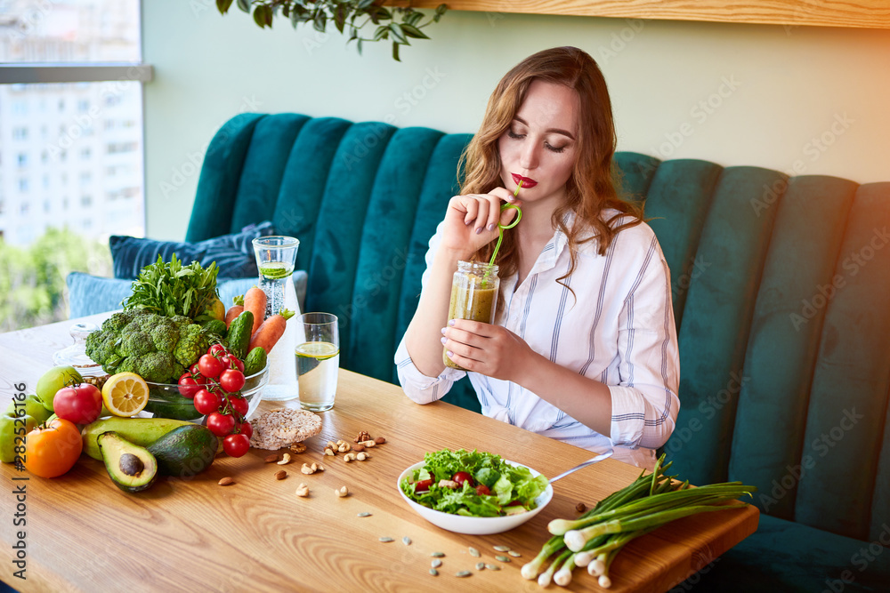 Plakat Young woman drinking smoothie in the beautiful interior with green flowers on the background and fresh fruits and vegetables. Healthy eating concept. Vegan meal and detox menu