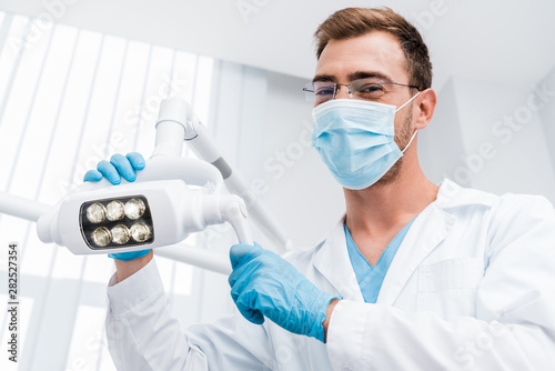 low angle view of dentist in glasses and medical mask touching medical lamp and looking at camera