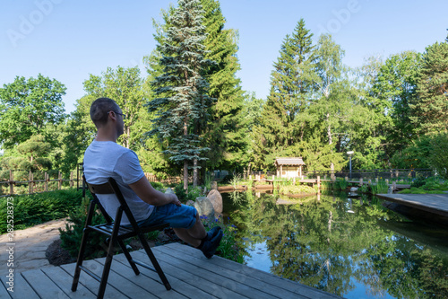 Young man meets dawn, sitting in front of a pond on a wooden folding chair, legs stretched forward