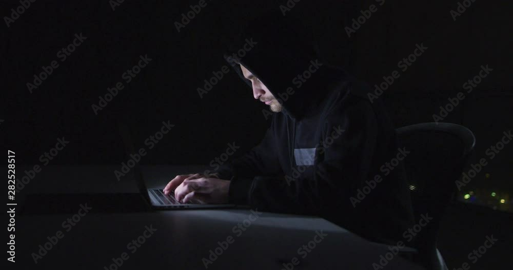 Hacker using laptop computer while working in dark office with big city light Hacker using laptop computer while working in dark office with big city light