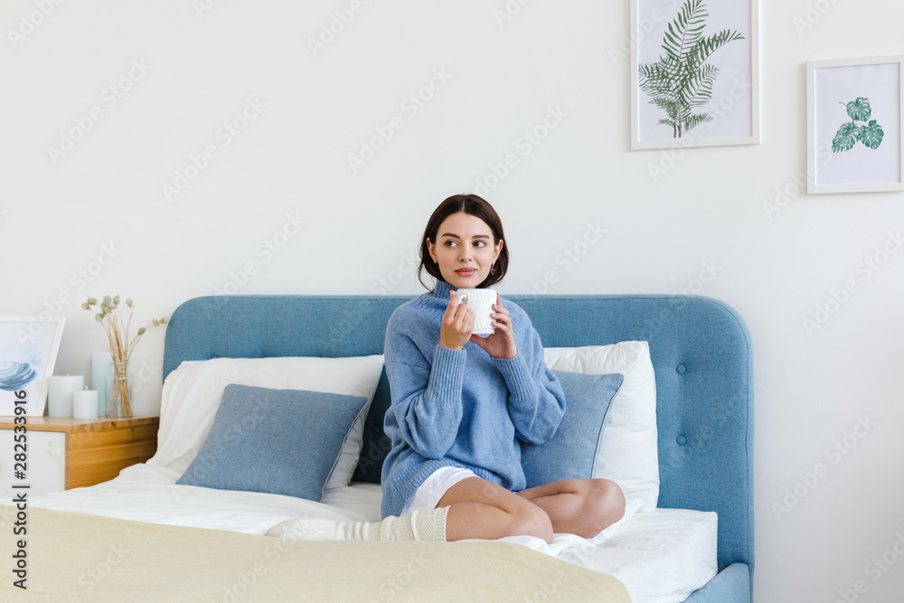 Obraz premium Dziewczyna w niebieskim swetrze we wnętrzu stylu Hygge z filiżanką w ręku siedzi na łóżku