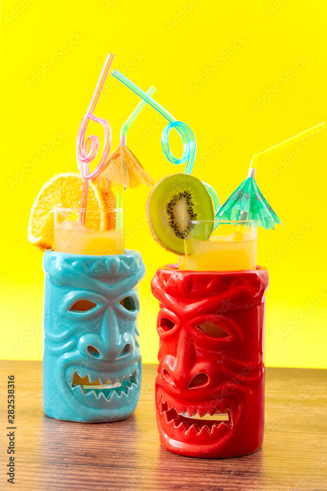 Hawaiian tiki bar, summer refreshing drinks and tropical cocktails concept theme with glasses of alcohol and juice mixed together, kiwi and orange slices, umbrella and drinking straw on wooden table