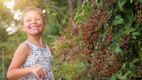 Authentic shot of a happy little girl with face smeared of berries is eating a fresh biologic blackberries just harvested by herself in a forest and smiling in the camera.