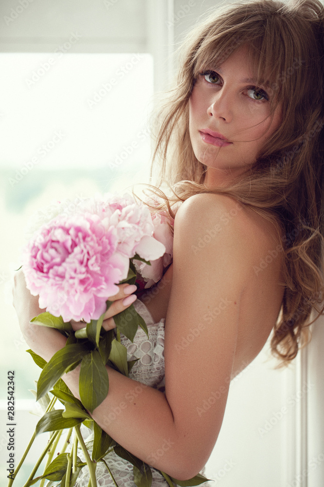 Beautiful blonde woman with sensual lips posing in lightfull white room among peonies flowers. - Image