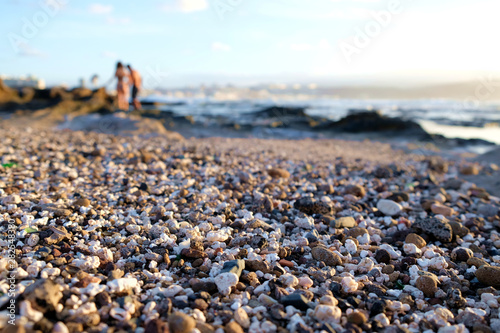 Canvas Print Pebbly beach in late afternoon with warm sunshine light and blurred human figures