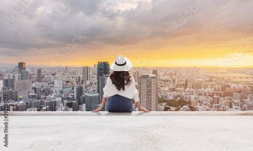 Woman in white hat and clothes sitting on rooftop floor, looking at Osaka city in sunset