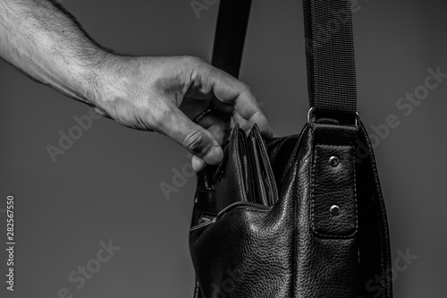 Black and white photo of a thief's hand pulling a purse from a leather bag on a gray background
