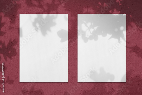 Two empty white vertical rectangle poster mockups with soft shadows on dark red colored concrete wall background. Flat lay, top view