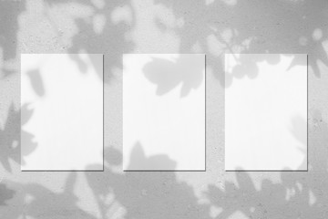 Three empty white vertical rectangle poster mockups with soft shadows on neutral light grey concrete wall background. Flat lay, top view