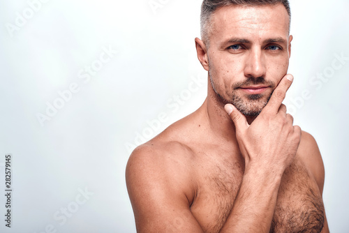 I am perfect. Handsome and sexy man with a stubble keeping hand on chin and looking at camera while standing against grey background
