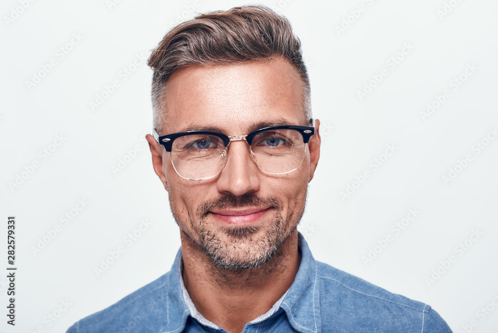 Happy and handsome. Close up portrait of charming bearded man in eyewear looking at camera and smiling while standing against grey background