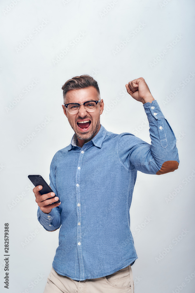 Success Portrait of handsome and happy bearded man in eyewear holding smart phone and keeping one hand clenched in fist while standing against grey background