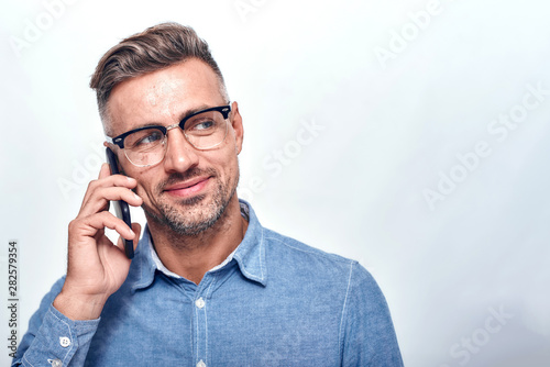 Glad to hear you Handsome and cheerful bearded man in casual wear talking on the phone and smiling while standing against grey background