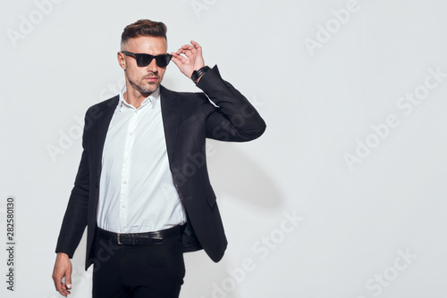 Confident and handsome. Good looking bearded businessman in classic suit adjusting eyewear and looking away while standing against grey background