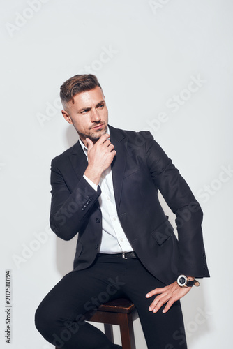 Smart and confident. Portrait of bearded charming businessman in classic suit looking at camera and holding hand on chin while sitting on the chair against grey background