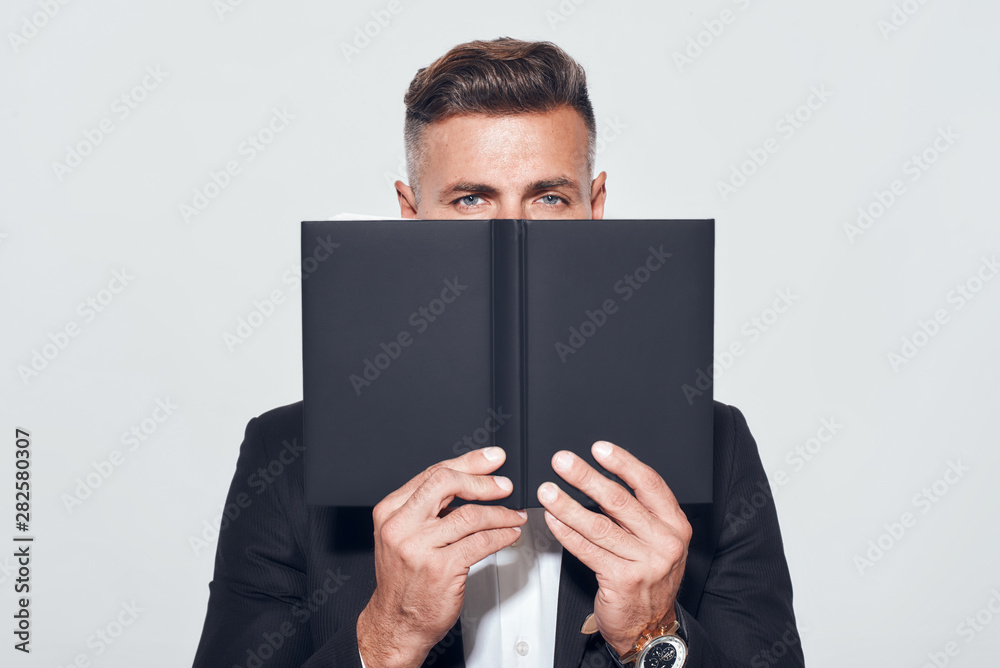 Professionalism. Confident businessman in classic wear covering his face with notebook and looking at camera while standing against grey background Professionalism. Confident businessman in classic wear covering his face with notebook and looking at camera while standing against grey background