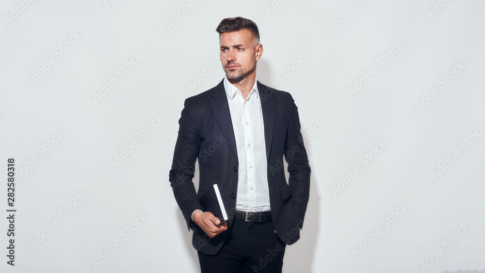 Ready to work. Stylish and confident businessman in classic wear holding a notebook and keeping hand in pocket while standing against grey background Ready to work. Stylish and confident businessman in classic wear holding a notebook and keeping hand in pocket while standing against grey background
