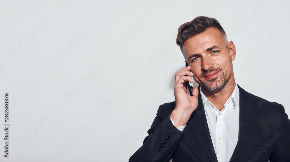 Good talk. Handsome bearded businessman talking on the phone and smiling while standing against grey background Good talk. Handsome bearded businessman talking on the phone and smiling while standing against grey background