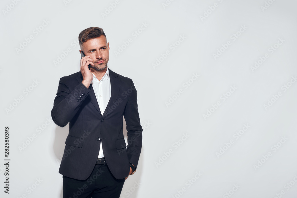 Talking about business. Handsome bearded businessman talking on the phone while standing against grey background Talking about business. Handsome bearded businessman talking on the phone while standing against grey background