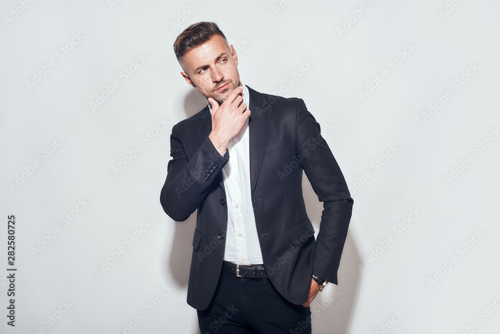 Handsome man. Bearded businessman in classic suit touching his chin and looking away while standing against grey background Handsome man. Bearded businessman in classic suit touching his chin and looking away while standing against grey background