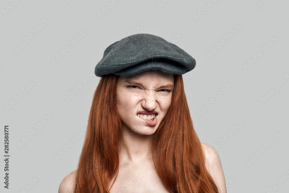 Rude girl. Portrait of young dissatisfied redhead woman in headgear biting a lip and looking at camera while standing against grey background Rude girl. Portrait of young dissatisfied redhead woman in headgear biting a lip and looking at camera while standing against grey background