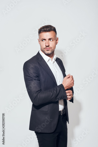 In his own style. Handsome bearded businessman in classic suit adjusting sleeves and looking away while standing against grey background