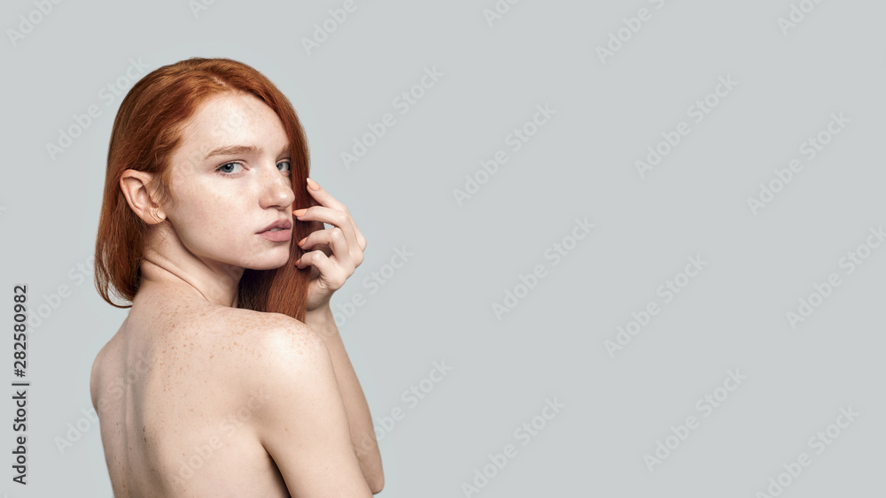 Feeling beautiful. Side view of young and gorgeous redhead woman looking at camera while standing against grey background Feeling beautiful. Side view of young and gorgeous redhead woman looking at camera while standing against grey background