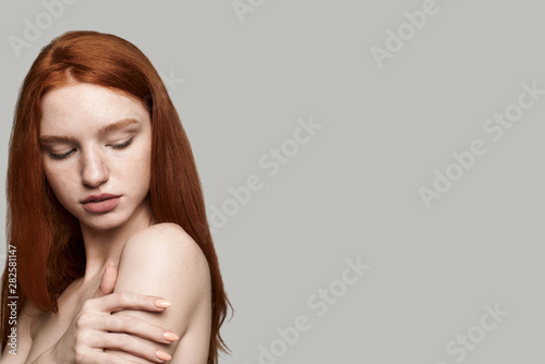 I love my skin. Beautiful young redhead woman touching her soft skin while standing against grey background