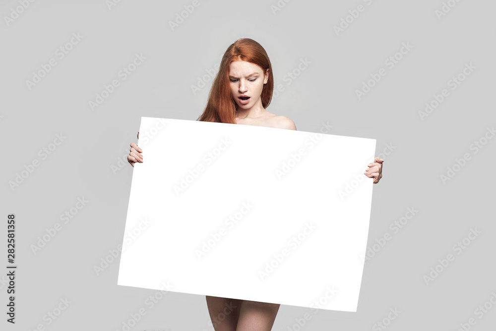 Shocked young redhead woman holding empty blank board and looking at it while standing against grey background Shocked young redhead woman holding empty blank board and looking at it while standing against grey background