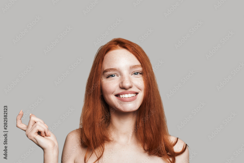Come here. Portrait of young and cute teenage girl with long red hair is gesturing and smiling while standing against grey background Come here. Portrait of young and cute teenage girl with long red hair is gesturing and smiling while standing against grey background
