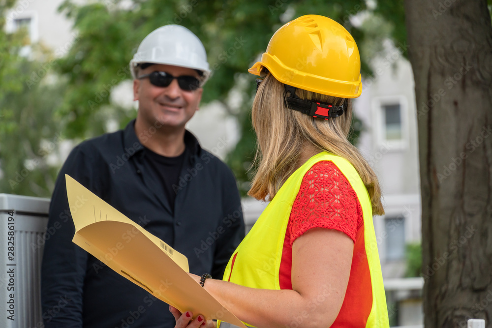 Back turned female construction worker with yellow safety jacket and hardhat. In the background, a male co-worker Back turned female construction worker with yellow safety jacket and hardhat. In the background, a male co-worker