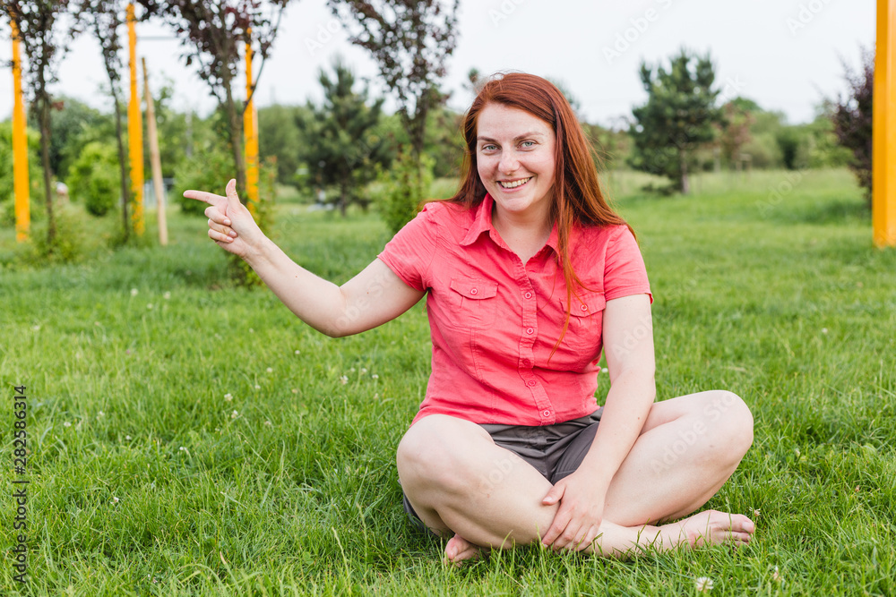 Image of beautiful cute young woman sitiing on grass in the park and pointing. Red-haired girl outdoors at a summer day Image of beautiful cute young woman sitiing on grass in the park and pointing. Red-haired girl outdoors at a summer day