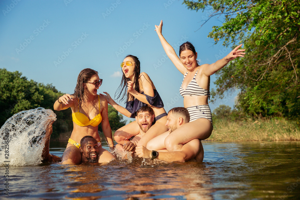 Obraz premium Happy group of friends having fun while laughting and swimming in river. Joyful men and women splashing water at riverside on the sunset in sun light. Summertime, friendship, resort, weekend concept.