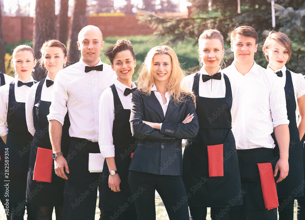 A large group of waiters and waitresses in the open air stand one after another
