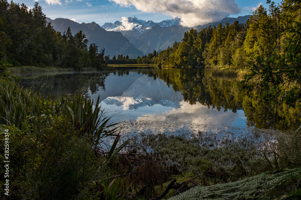 A landscape view of the incredibly beautiful Lake Matheson, New Zealand with the reflection of the stunning Southern Alps and the majestic Mt Cook in the still waters, foliage in the foreground