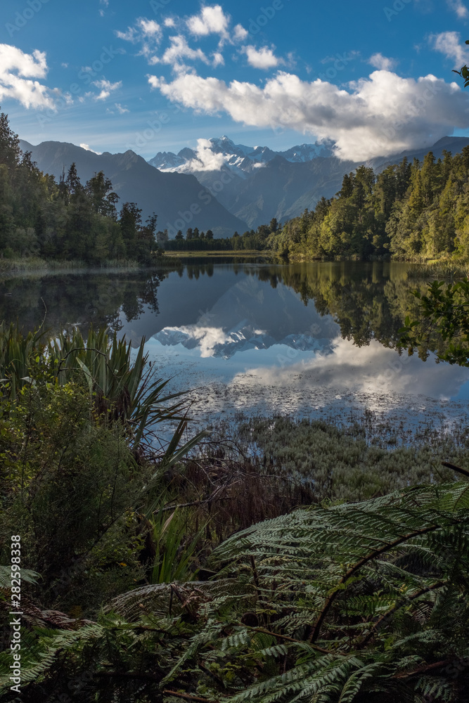 A portrait view of the incredibly beautiful Lake Matheson, New Zealand with the reflection of the stunning Southern Alps and the majestic Mt Cook in the still waters, foliage in the foreground