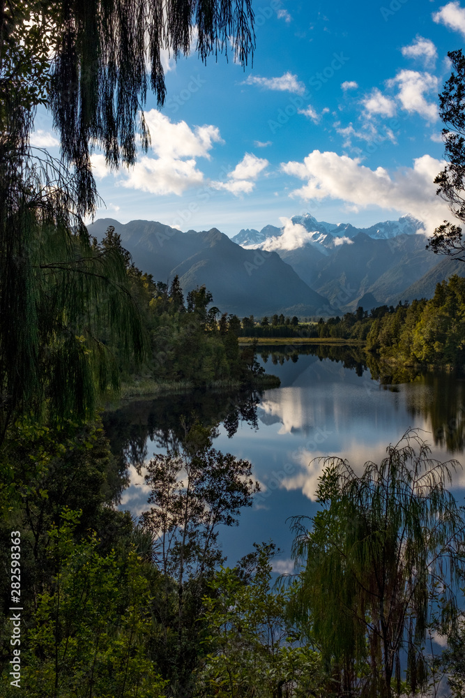 A portrait view framed by foliage of the incredibly beautiful Lake Matheson, New Zealand with the reflection of the stunning Southern Alps and the majestic Mt Cook in the still waters.
