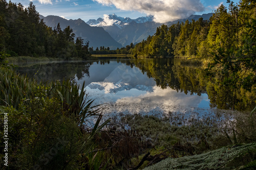 A landscape view of the incredibly beautiful Lake Matheson, New Zealand with the reflection of the stunning Southern Alps and the majestic Mt Cook in the still waters, foliage in the foreground