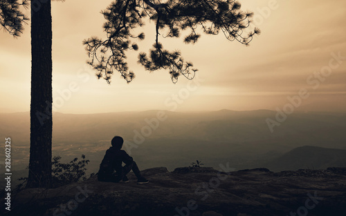 Silhouette of alone man sitting under big tree on the mountain view with fog in sunset background. Backpacker travel lifestyle and sad concept.