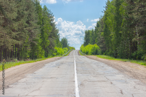 A poorly-paved rural two-lane road, deciduous forest, cloudy sky, highway to the horizon