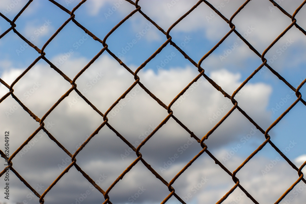 Old rusty mesh netting on a blue sky with clouds, background wallpaper texture.