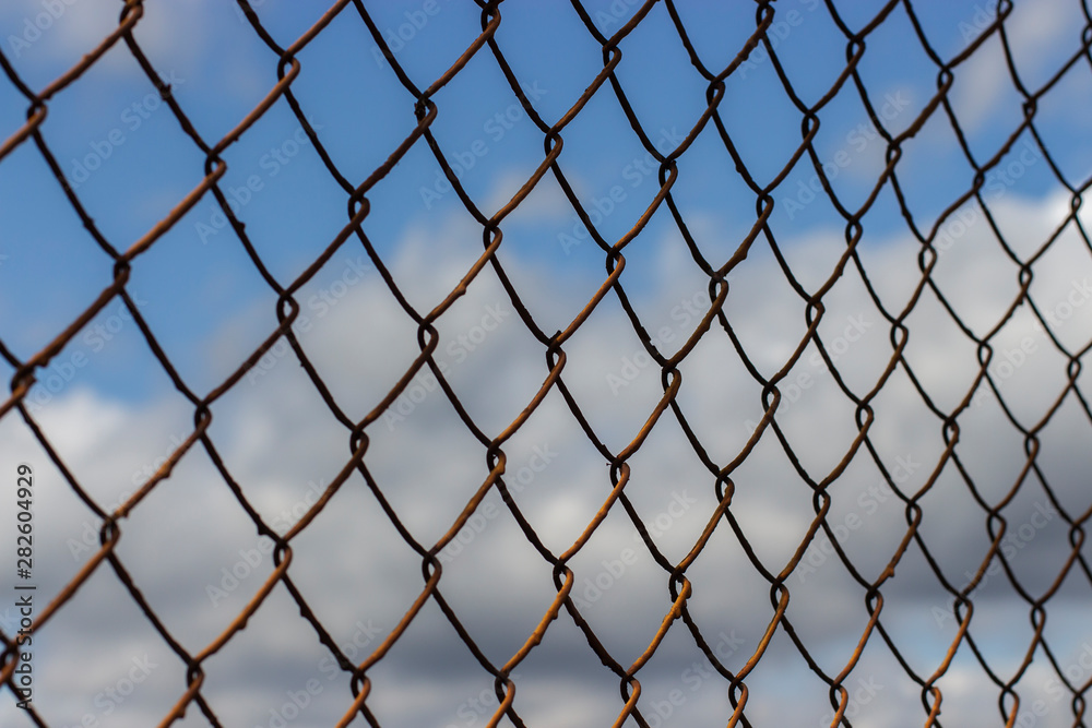 Fototapeta premium Old rusty mesh netting diagonally against a blue sky with clouds, background wallpaper texture
