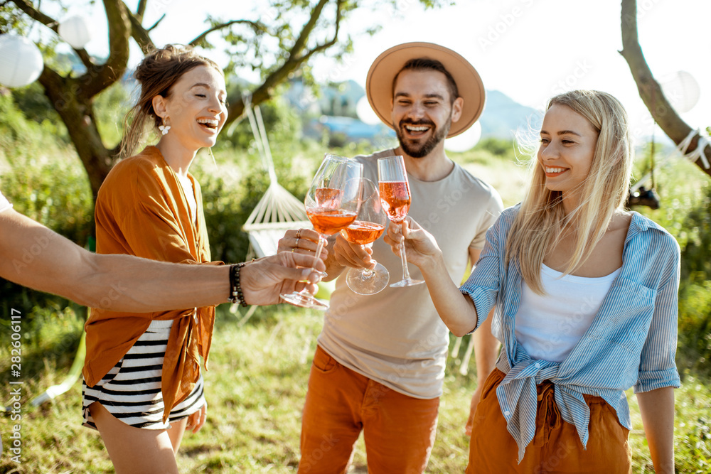 Fototapeta premium Young and cheerful friends having fun, clinking with wine glasses in the beautifully decorated backyard during a festive meeting or party on a sunny summer evening