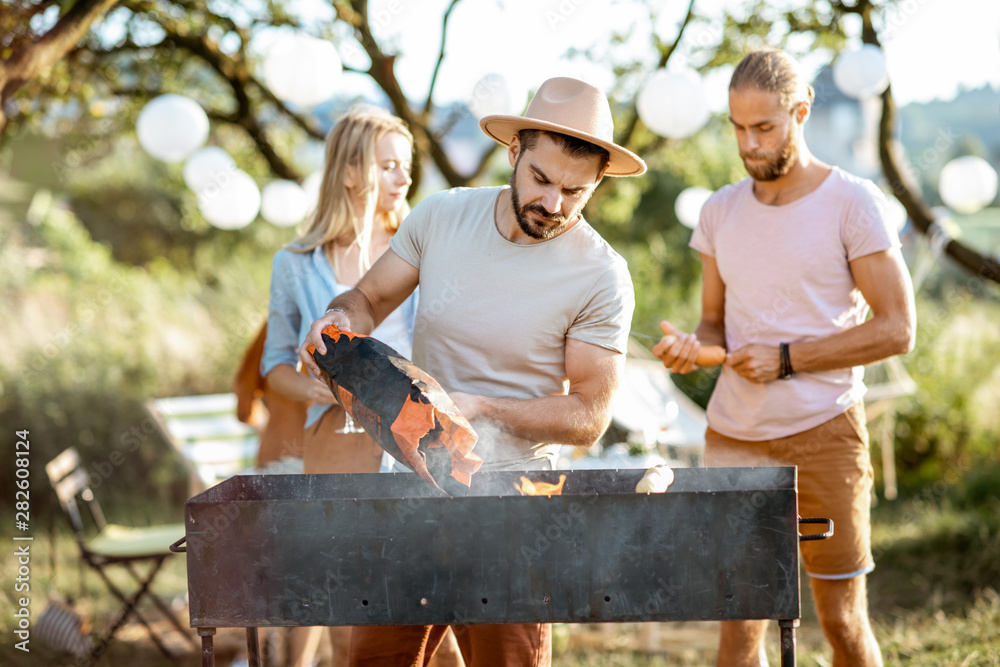 Obraz premium Group of a young friends having barbecue, cooking food in the beautiful garden during a festive lunch on a summer aftenoon